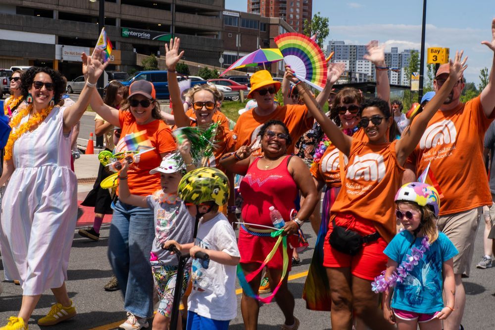 A group of smiling New Democrat MLAs wave and cheer as they march together in a Pride parade. Rainbow flags, leis, and orange shirts are visible, with children joining in on scooters.