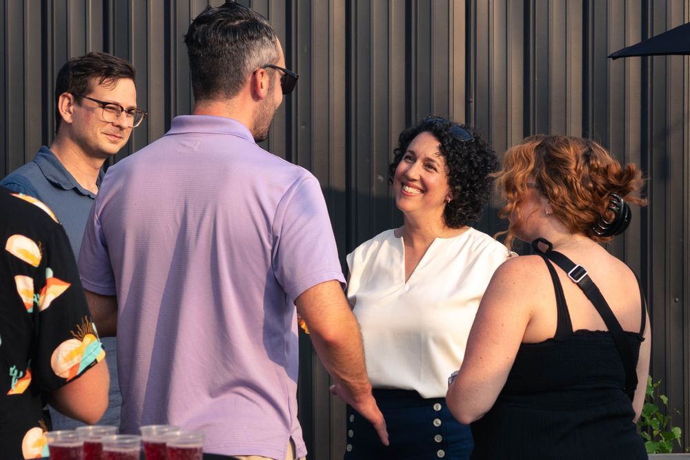 Claudia Chender wearing a white blouse smiles warmly while talking with three people outdoors near a dark building. Drinks sit on a table in the foreground.