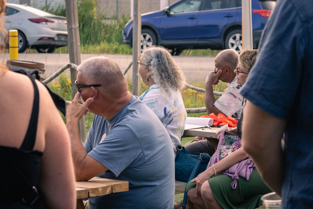 Several people sit at picnic tables outdoors, listening attentively. Cars are parked in the background near a fence.