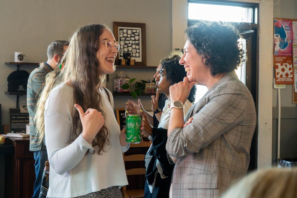 Claudia and a Cape Breton woman laugh and engage in an animated conversation at a community gathering. One holds a green Bubly can, and the background shows more attendees talking and smiling.