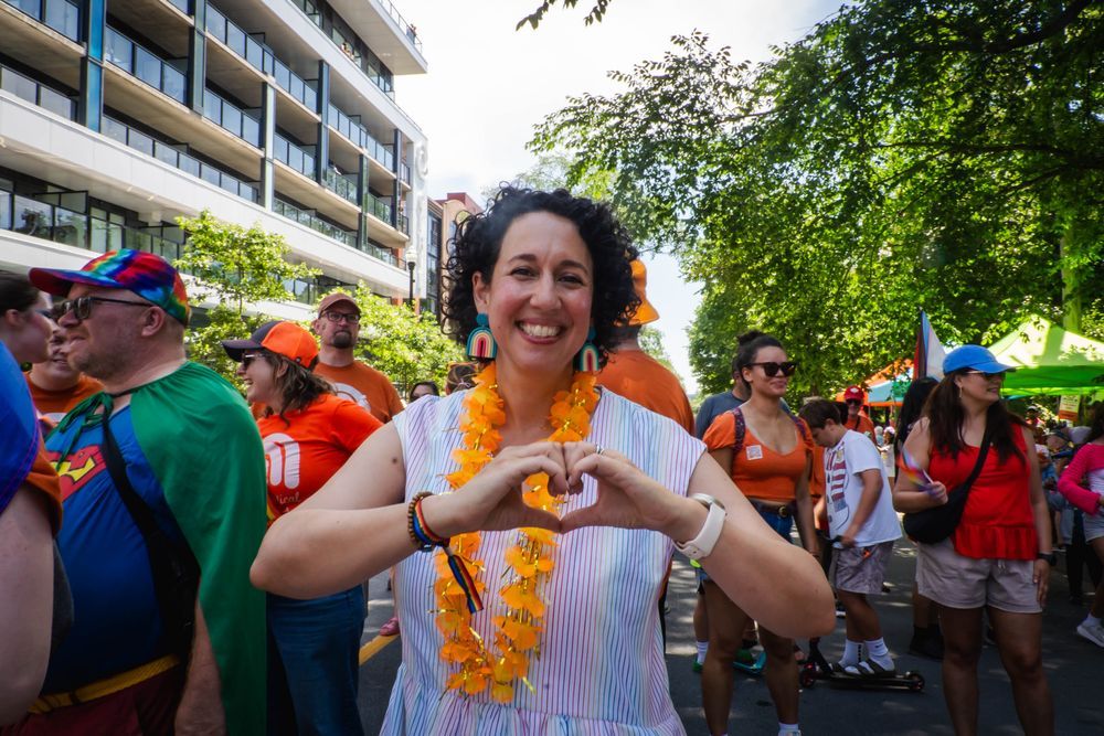 Claudia Chender in striped sleeveless dress and rainbow earrings stands in the middle of a lively Pride parade, forming a heart shape with her hands. Orange leis and a crowd in colourful outfits fill the background.
