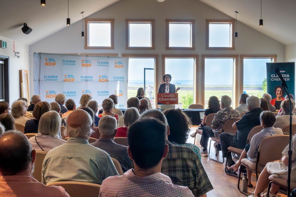 A large audience sits in rows as Nova Scotia NDP Leader Claudia Chender speaks at a podium in a bright, high-ceilinged room with NSNDP banners and a backdrop with her name. A vertical sign with her photo stands to the right.