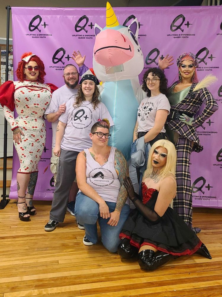 Staff members of Q Plus, wearing Q Plus staff tshirts, pose with Q Plus' unicorn mascot Tapioca, and drag performers Mia E. Z'lay, Ambrosia Black, and Miss Kiki Lucia in front of a purple Q Plus backdrop at The Firestone in Manchester, CT.