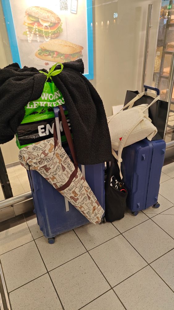 A picture showing all the luggage I have with me for cons. From left to right you see a big blue suitcase, with a shopper + coat on top of it, and an extra bag with my poster rack in it hanging off the handle. Next to it is my personal (black) tote bag on the floor, with a medium blue suitcase on the other side. On top of that is a white cooling backpack with all the food items, and lastly a black paper bag with some goodies bought at the con. The picture was taken on the side of the Albert Heijn To Go on Schiphol.