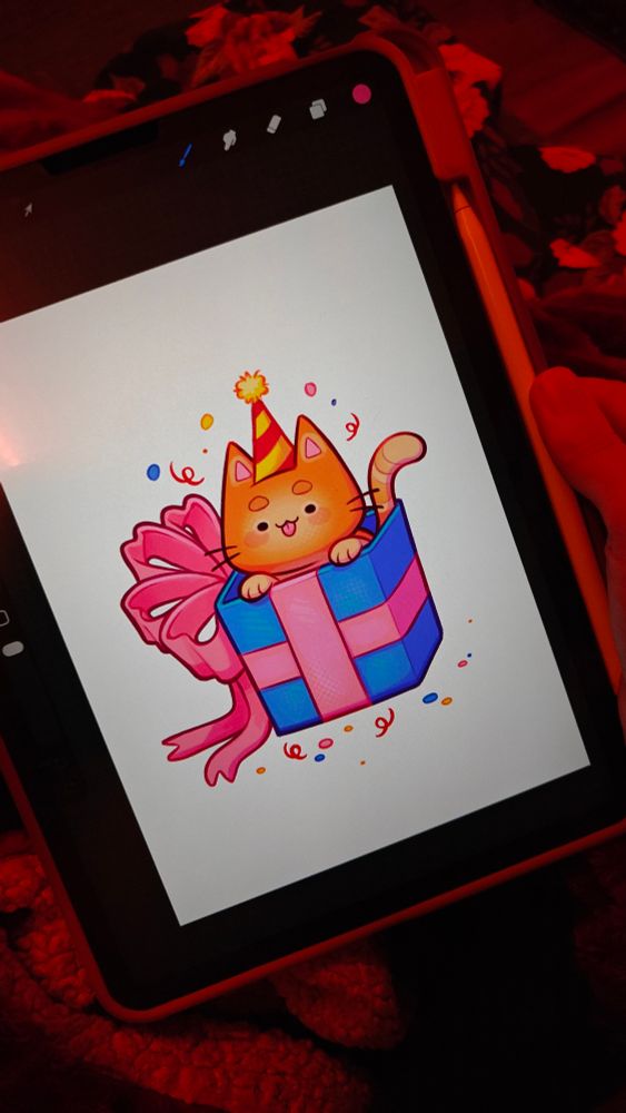 A picture of my iPad. On it it's showing a(n almost) finished drawing of a kitty with a little birthday hat, peeking out of a box with a big ribbon around it. The drawing seems to be already fully rendered, though I might still work on it/change some things later.