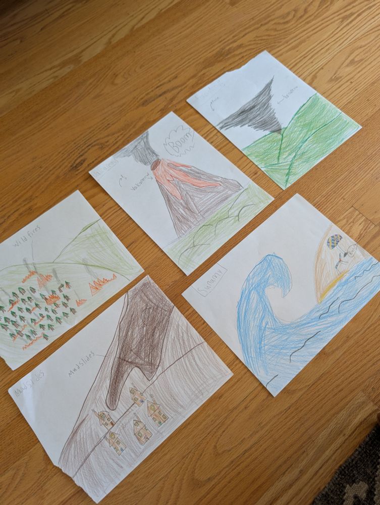 Six children's drawings on white paper are laid out on a wooden floor. Each drawing depicts a natural disaster. From top left to bottom right, the drawings show: a forest with flames labeled "wild fires"; a volcano erupting with lava labeled "volcanoes" and "Boom"; a tornado over green hills labeled "tornadoes"; a hillside with trees on fire labeled "wild fires"; a large mudslide heading toward houses labeled "mud slides"; and a giant wave approaching a coastline labeled "tsunami". The drawings are colorful and done with crayons or colored pencils.
