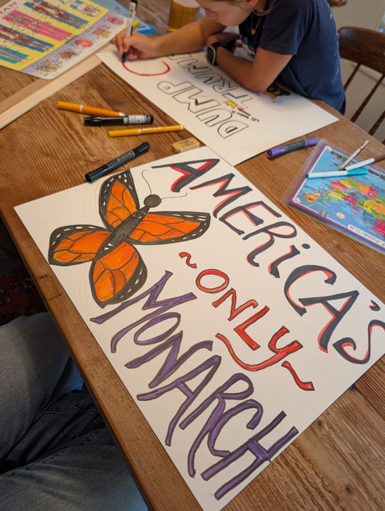 Two people are making protest signs at a wooden table. One sign in the foreground reads “AMERICA’S ONLY MONARCH” with a large, brightly colored monarch butterfly illustration, using red and purple lettering. In the background, another person is working on a sign that says “DUMP TRUMP!” with a red circle and line crossing out the name. Various markers, a U.S. map, and a butterfly identification guide are also on the table.
