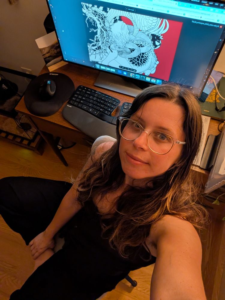 A woman with long wavy brown hair and clear-framed glasses is sitting cross-legged on a chair, smiling slightly as she takes a selfie from above. She is wearing a black sleeveless outfit. Behind her is a wooden desk with a large monitor displaying a black, white, and red illustration of a woman intertwined with a dragon. A keyboard, ergonomic mouse, and various papers are also visible on the desk. The lighting is warm, giving the room a cozy feel.
