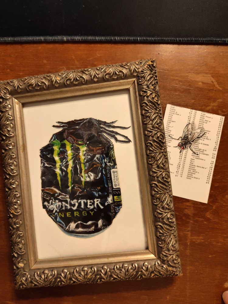Small oil painting of a crushed Mint energy can with a small crab resting at the top, displayed in an ornate antique silver carved frame. A tiny painting of a fly on a piece of a page of map index sits beside it. 