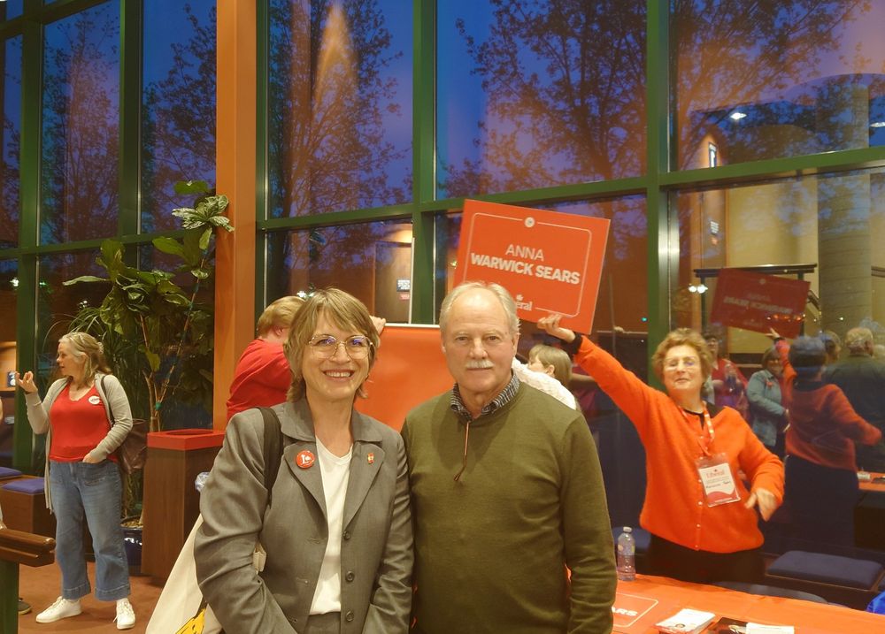 Anna Warwick Sears, the Liberal candidate for Vernon-Lake Country-Monashee, poses smiling with Vernon Mayor Victor Cumming at an indoor campaign event. Behind them, a supporter holds up a red sign with Anna's name on it. Other people wearing red attire are visible in the background, including one woman wearing a name badge and gesturing enthusiastically. The event takes place in a well-lit building with large windows revealing a twilight sky and reflections of trees.

