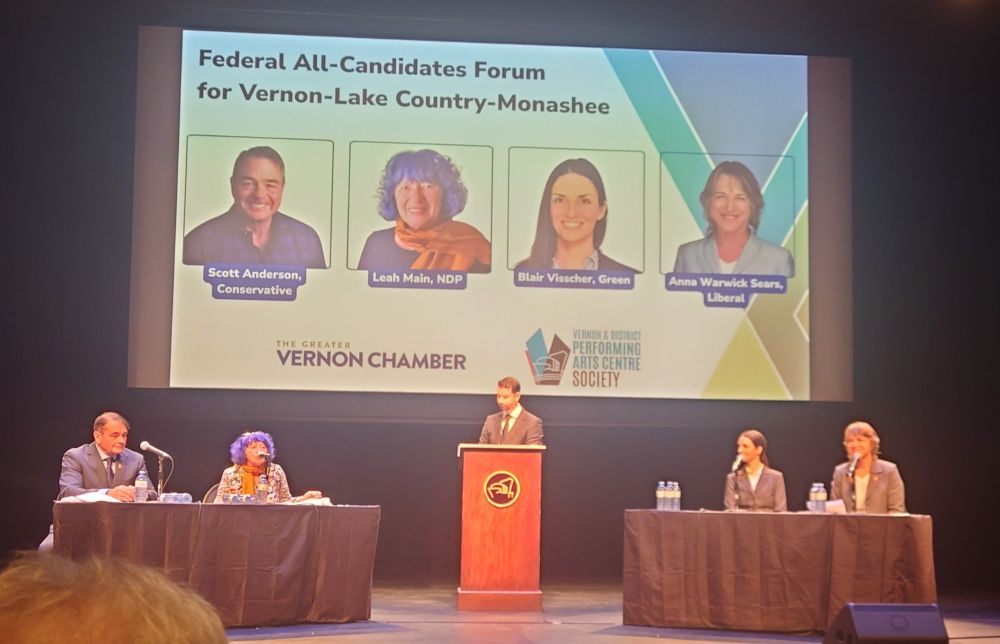 A photo of a federal all-candidates forum for Vernon-Lake Country-Monashee, held on a stage at the Vernon & District Performing Arts Centre. A large screen above shows headshots and names of four candidates: Scott Anderson (Conservative), Leah Main (NDP), Blair Visscher (Green), and Anna Warwick Sears (Liberal). The event is hosted by the Greater Vernon Chamber. On stage, Scott Anderson and Leah Main are seated at a table on the left, the moderator stands at a podium in the center, and Blair Visscher and Anna Warwick Sears are seated at a table on the right.

