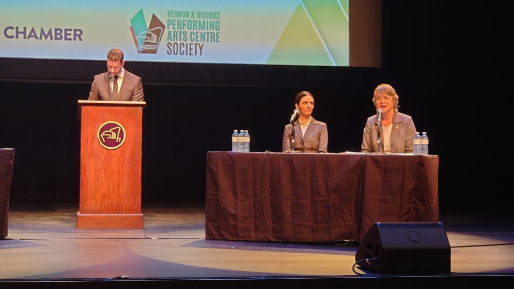 
At the federal all-candidates forum for Vernon-Lake Country-Monashee, two candidates—Blair Visscher (Green) and Anna Warwick Sears (Liberal)—are seated at a table on stage. Anna is speaking into a microphone, while Blair listens attentively. The moderator stands at a wooden podium to the left, looking down at his notes. A logo for the Vernon & District Performing Arts Centre Society is displayed on the screen behind them. Multiple water bottles are on the table in front of each candidate.

