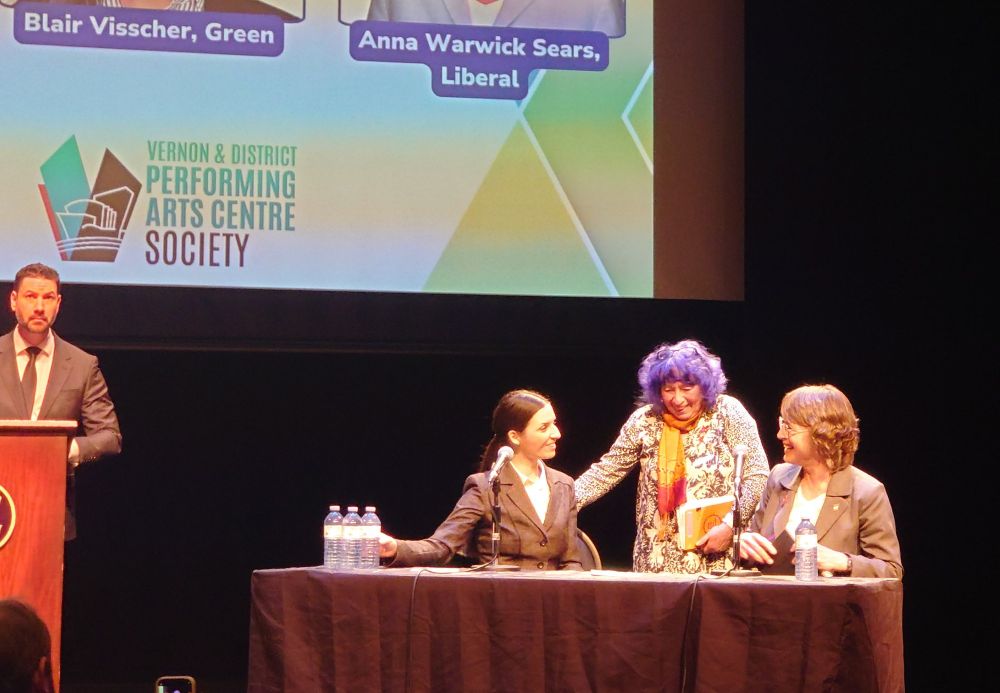 
During the federal all-candidates forum for Vernon-Lake Country-Monashee, three women share a warm moment on stage. Leah Main (NDP) stands smiling between Blair Visscher (Green), who is seated and looking up at her, and Anna Warwick Sears (Liberal), who is also seated and smiling. They appear to be exchanging friendly words after the event. The moderator stands at a podium to the left. The backdrop displays the Vernon & District Performing Arts Centre Society logo and names of the candidates. Bottles of water are on the table in front of the candidates.

