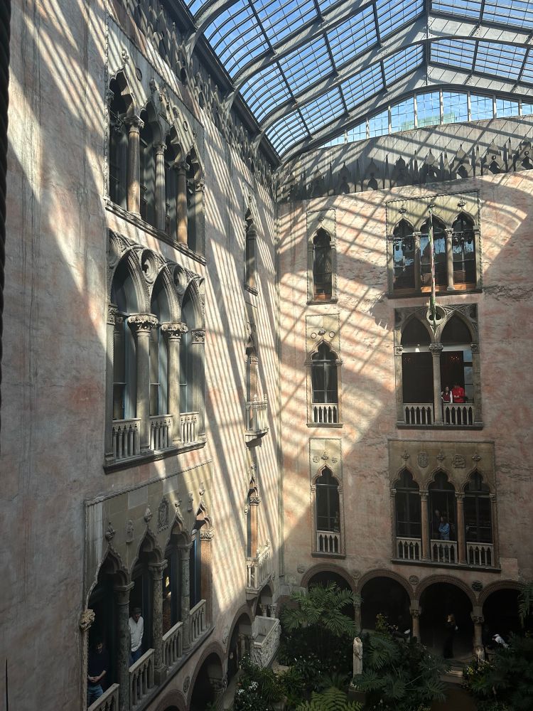 Photo of the courtyard/atrium of the Isabella Stewart Gardner Museum in Boston. The walls are a warm stucco, with ornate arched windows spanning three stories, with a glass roof that lets in natural light, which falls in a dramatic triangle over the walls. 