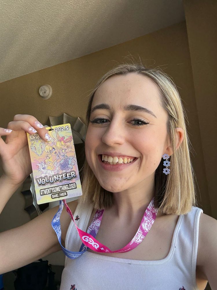 Lee (woman with blonde bob, white tank and blue flower earrings) is smiling and wearing her Games Done Quick name tag and lanyard