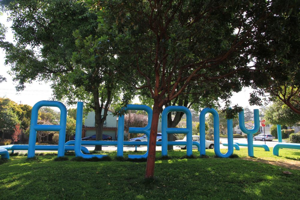 A blue large sculpture depicting the letters A, L, B, A, N, and Y, sits along a leafy bike path in Albany, California. 