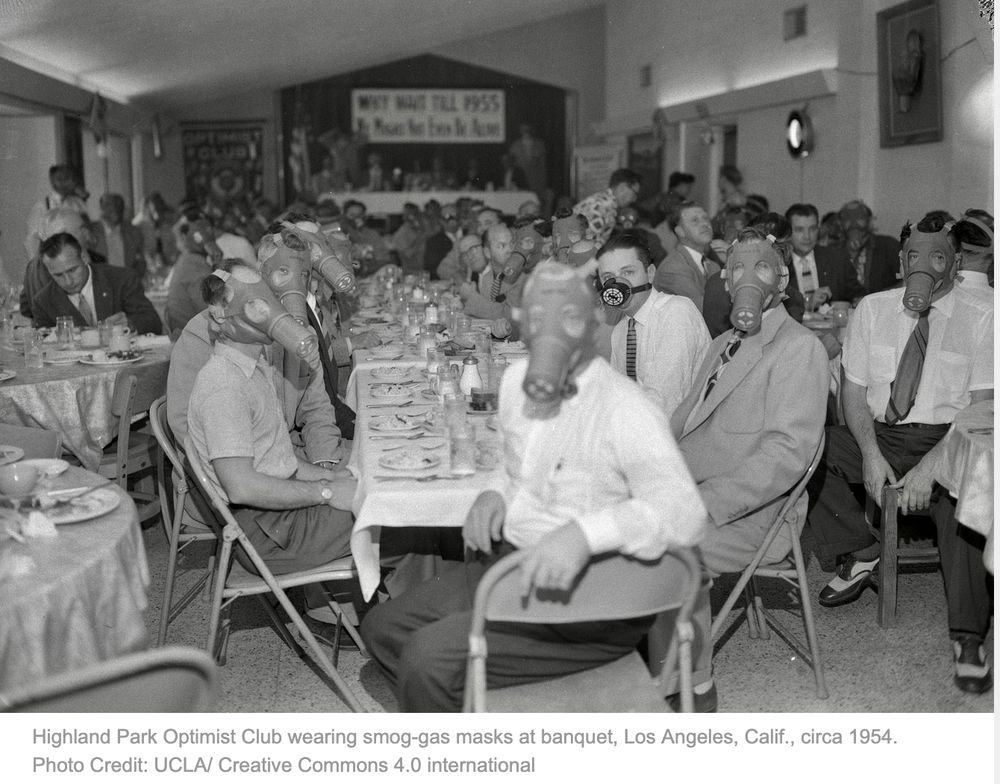 A group of men sit around a large table wearing what look like gas masks. The caption reads: Highland Park Optimist Club wearing smog-gas masks at banquet, Los Angeles, Calif., circa 1954