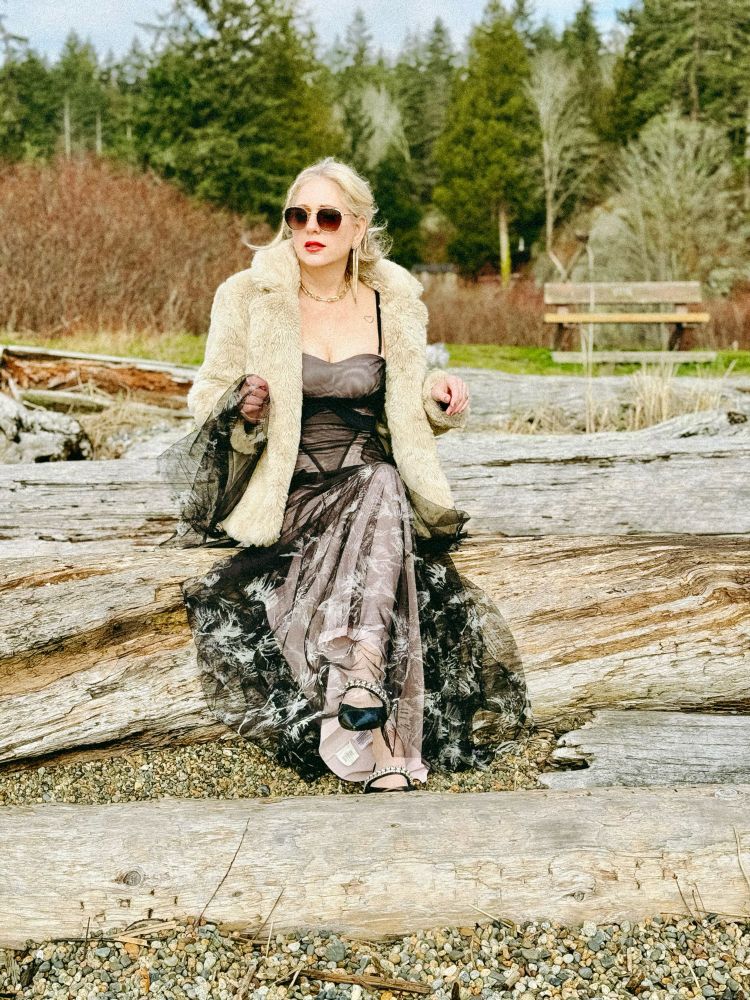 Sitting on a log in a light coloured faux fur and black and grey ballgown. Sunglasses on.