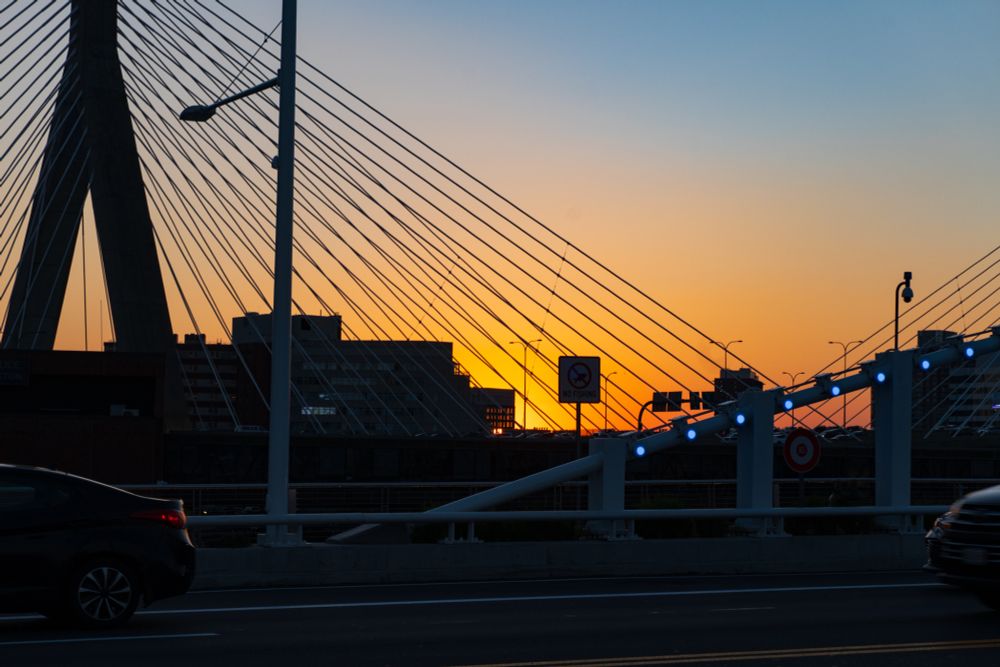 Sunset at Bunker Hill Memorial Bridge, shot from the nearby Bill Russell bridge, with lots of vehicle traffic present.