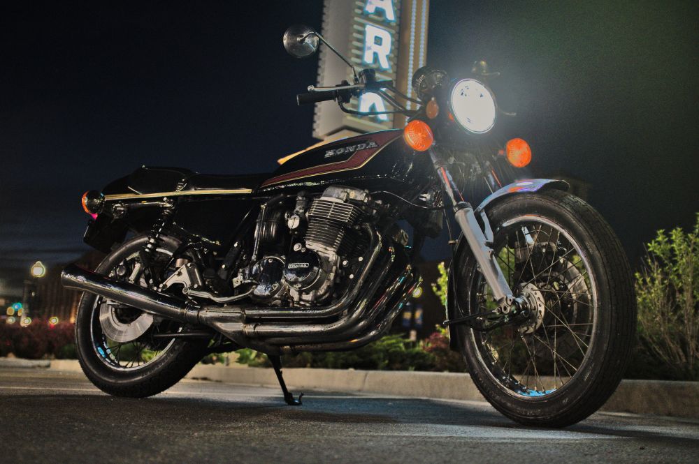 A Honda CB750 parked in front of a blue neon 'PARK' sign at night. The bike's headlight and side marker lights are turned on.