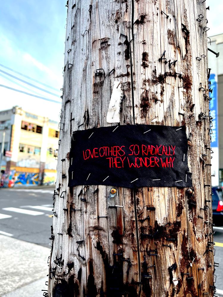 A scrap of black fabric is stapled to a utility pole with the words “Love others so radically they wonder why” embroidered with red thread.