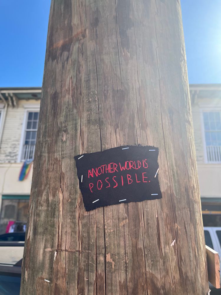 A scrap of black fabric is stapled to a utility pole with the words “Another world is possible “ stitched with red thread. Blue sky and a building are visible in the background.
