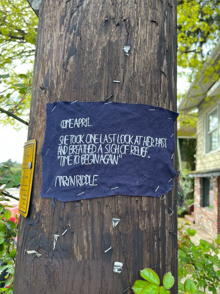 A scrap of dark blue fabric is stapled to a utility pole with the word:

COME APRIL

she took one last look at her past and breathed a sigh of relief.

“time to begin again”

/taryn riddle
stitched with white thread. Trees and road are visible in the background 