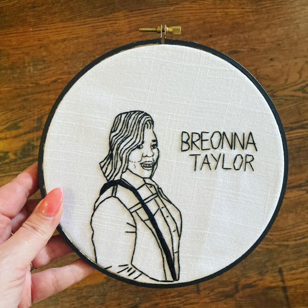 I’m holding a finished embroidery hoop with a stitched portrait of Breonna Taylor, a young black woman with shoulder length hair, smiling. The embroidery was done with black thread on white and her name is stitched next to her.