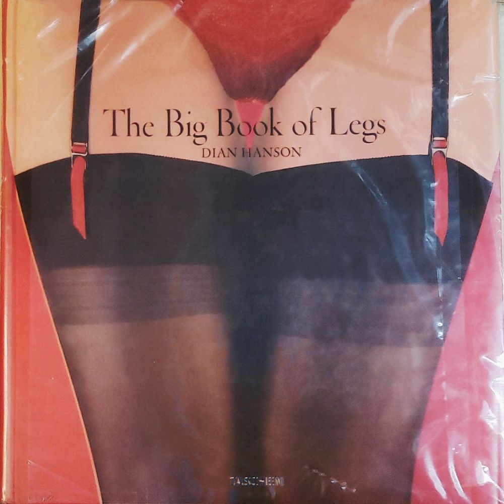 A photo of "The Big Book of Legs" by Dian Hanson, published by Taschen. The image is a woman's legs from hips to knees, wearing lacy red panties and black gartered stockings with red ribbons. The cover is wrapped in a custom plastic sleeve to protect it's clear plastic dustcover, which has begun to crack at the spine. The lingerie in the photo is printed on the removable clear cover. Concealed by the clear cover is the same photo, without the lingerie.