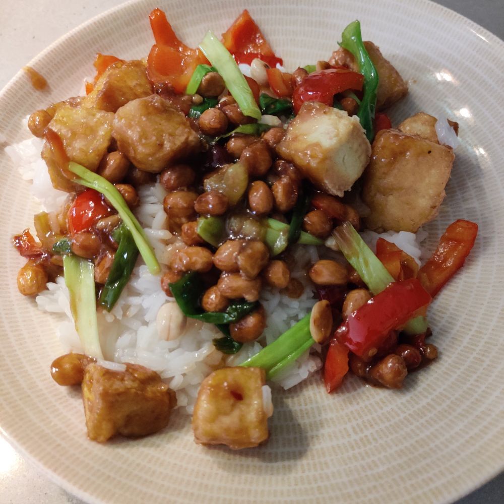 Beigellä lautasella riisiä, tofua kung pao -kastikkeessa, paahdettuja maapähkinöitä, paprikaa ja kevätsipulia