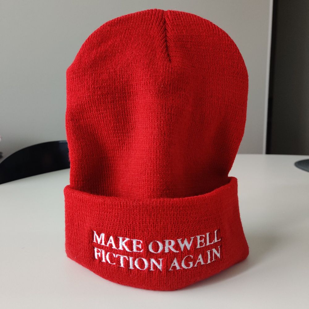 punainen pipo johon on kirjailtu valkoisella teksti "Make Orwell fiction again"