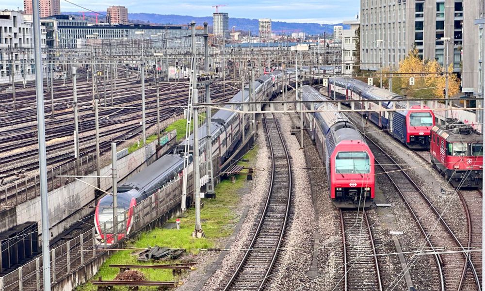 A train pulls into the Zurich Museumstrasse train station, next to a few other s-Bahn trainsets parked on a siding, with the track field before Zurich main station in the background.