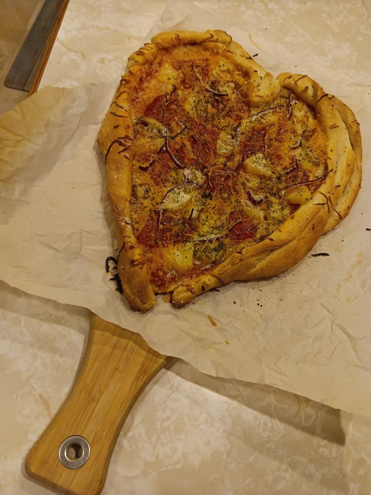Heart shaped pizza on parchment paper with the handle of a charcuterie board poking out. It sits on a counter. 