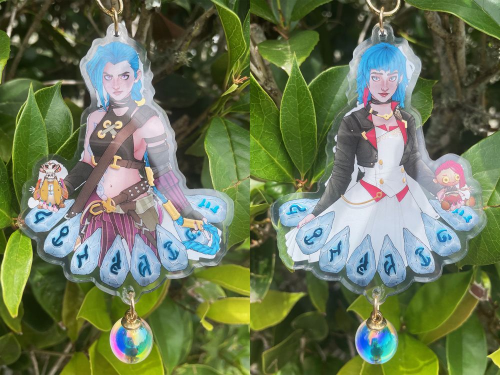 A double sided charm of Jinx from Arcane. The front shows Jinx’s regular appearance holding her monkey bomb, while the back shows the alternate universe version of Powder from Arcane S2E7 holding the doll of Vi from her memory shrine.