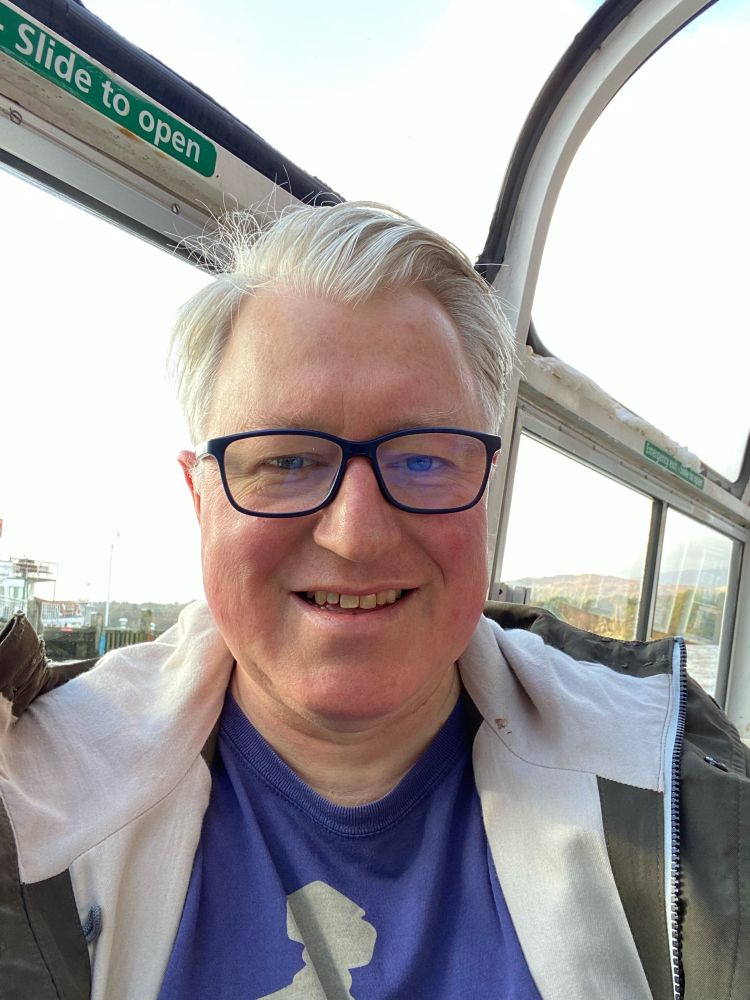 Selfie of myself. On a boat I have a green coat blue T shirt, black glasses, grey hair, parted on the left side