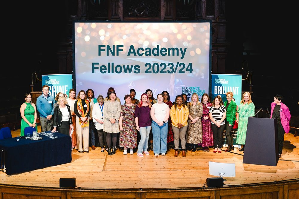 A group of around 25 people standing on a stage smiling at the camera. They have a large scree behind them saying FNF Academy Fellows 2023/34 and there are two pull up banners, on on either side of the screen. There is a table on the left and a lectern on the right of the stage.