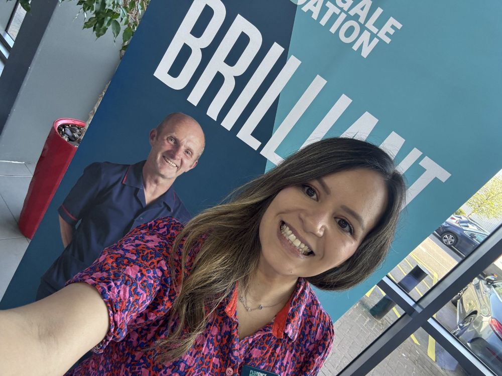 Image of a woman with brown hair in a floral top smiling, the photo is slanted to the right. There is a pull up banner with a man in nurses uniform in the background and the work brilliant above his head.