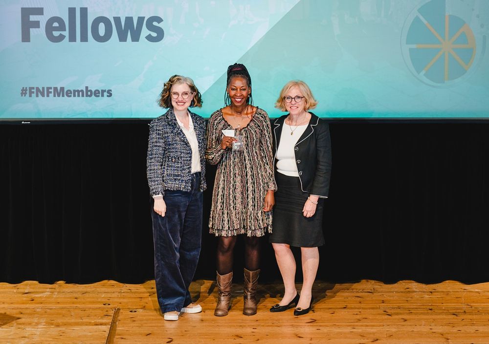 Three women on a wooden floor, smiling. The woman in the middle is holding a small bag with a badge inside. There is a screen behind with the word Fellows top left and beneath that there is the hashtag #FNFMembers.