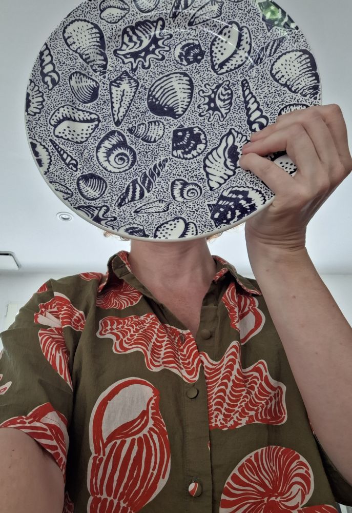 Selfie avec assiette bleu motifs coquillages devant ma tête. Je porte une chemise kaki à motifs coquillages rouge et blanc