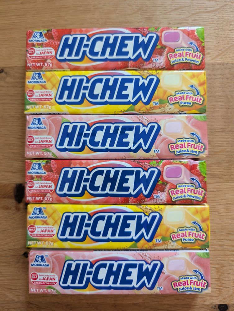 6 untereinander liegende längliche packungen süßigkeiten mit dem namen "hi-chew". in den gesckmacksrichtungen erdbeere, zitrone, pfirsich, erdbeere zitrone, pfirsich.
