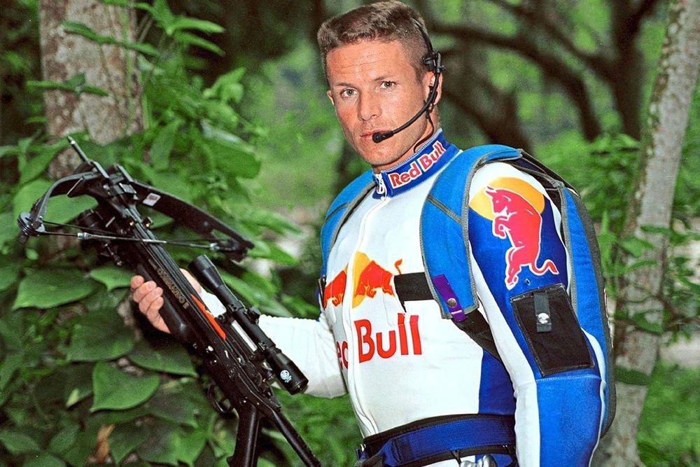 junger felix baumgartner seht vor einem wald er hat einen aerodynamischen red bull sportanzug (vllt zum skydiven oder so) an an und trägt einen blauen rucksack, am kopf hat er so ein headset wo mikro vom hor zum mund geht, so callcenter mäßig. er schaut in die karmera in der rechten hand hält er eine sportarmbrust