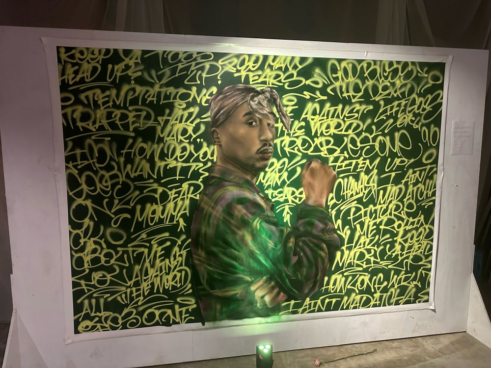 Photo of a painting of Tupac.  He’s wearing green.  The background is lyrics to his songs painted in yellow.