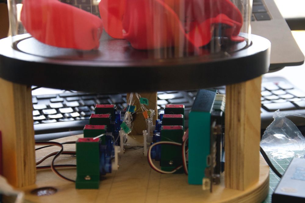 Side view of the servos mounted inside of the dome platform with fish line attached all going up into the hole where there aluminum rod stem has been fitted. Each fish line is labeled with their petal/servo number using various colored washi tape. There is a bottom view of the fallen petals on top of the dome base with the clear plastic dome cover on.  A black keyboard is in the background behind this enchanted rose project.