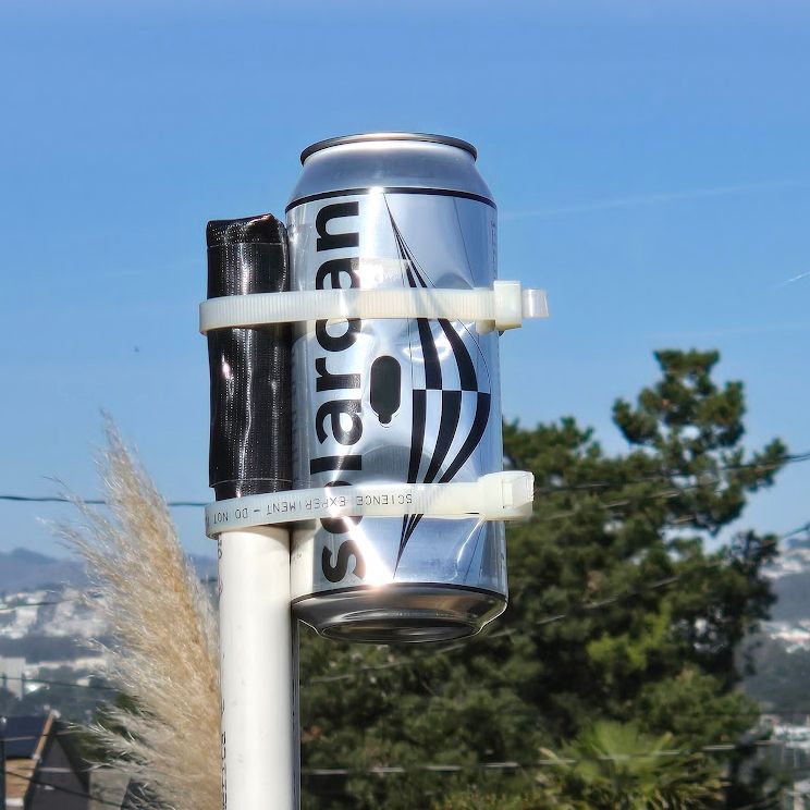 Aluminum can with words "solarcan" printed on the side and mounted with 2 big white cable ties to pvc pipes that were mounted to fence. Background is bright clear blue skies and green tree.