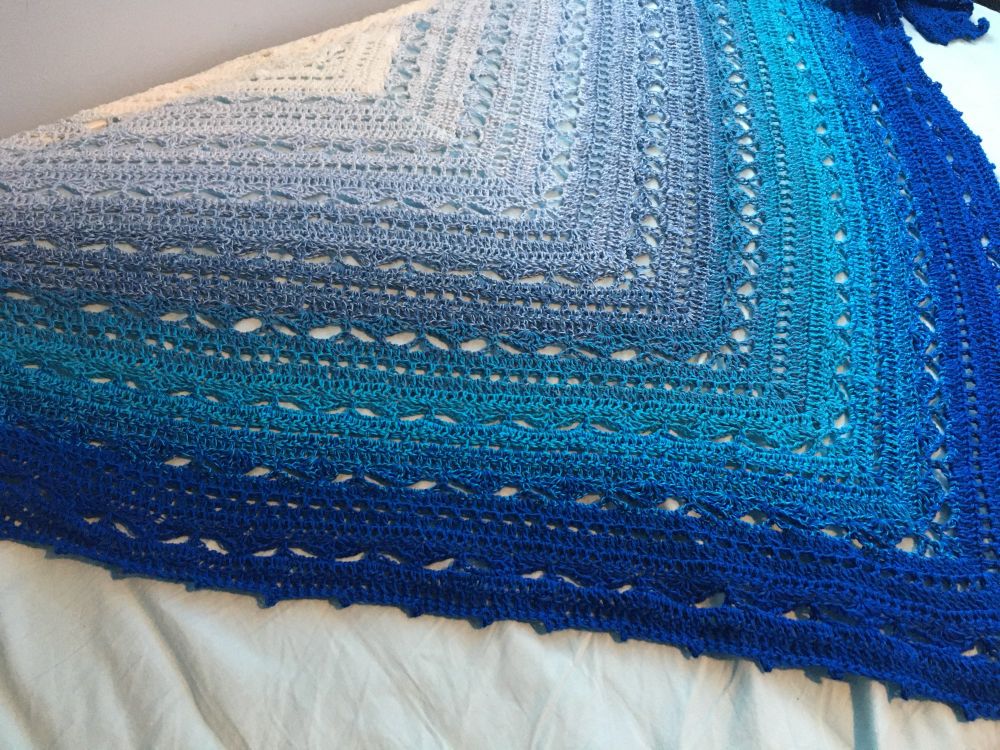 Bruinen triangle shawl by Morben Design. Starting in white at the top and gradienting through shades of blue from light to dark. 

In homage to the Water of Rivendell, the river Bruinen protected Frodo from the Nazgul. There is an Evenstar jewel design that runs down the middle. 

Cotton-acrylic. 