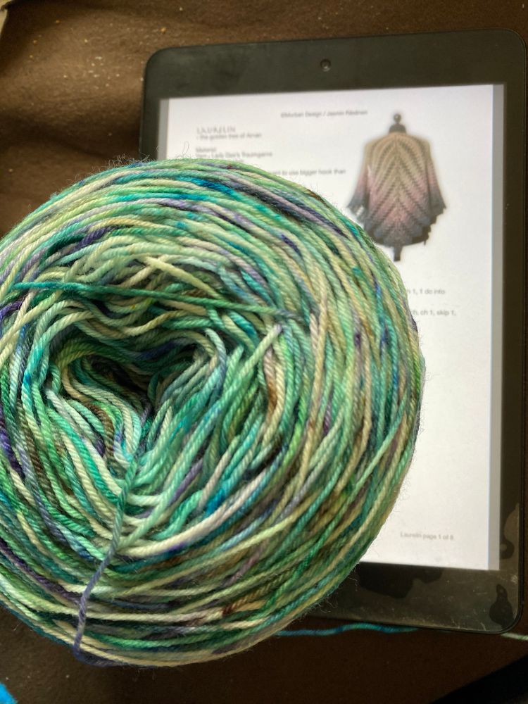 A cake of merino yarn in greens, blues, and teal with speckles of white and purple. It’s sitting on an iPad showing the title of the pattern: Laurelin