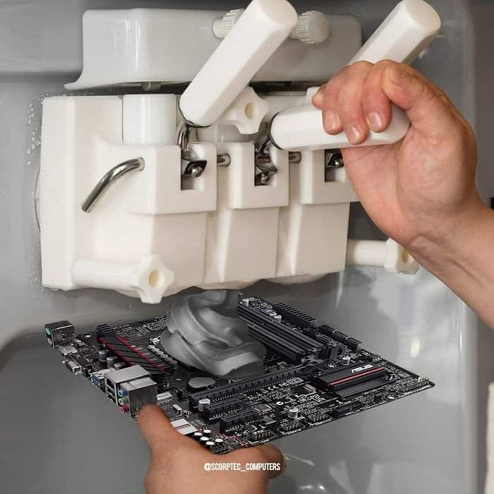 A PC motherboard with a dark grey ice cream looking substance being dispensed onto it by an ice cream machine. The substance is the colour of thermal paste. It is being deposited onto the CPU area and there is a dollop about the size of a tennis ball on it.
