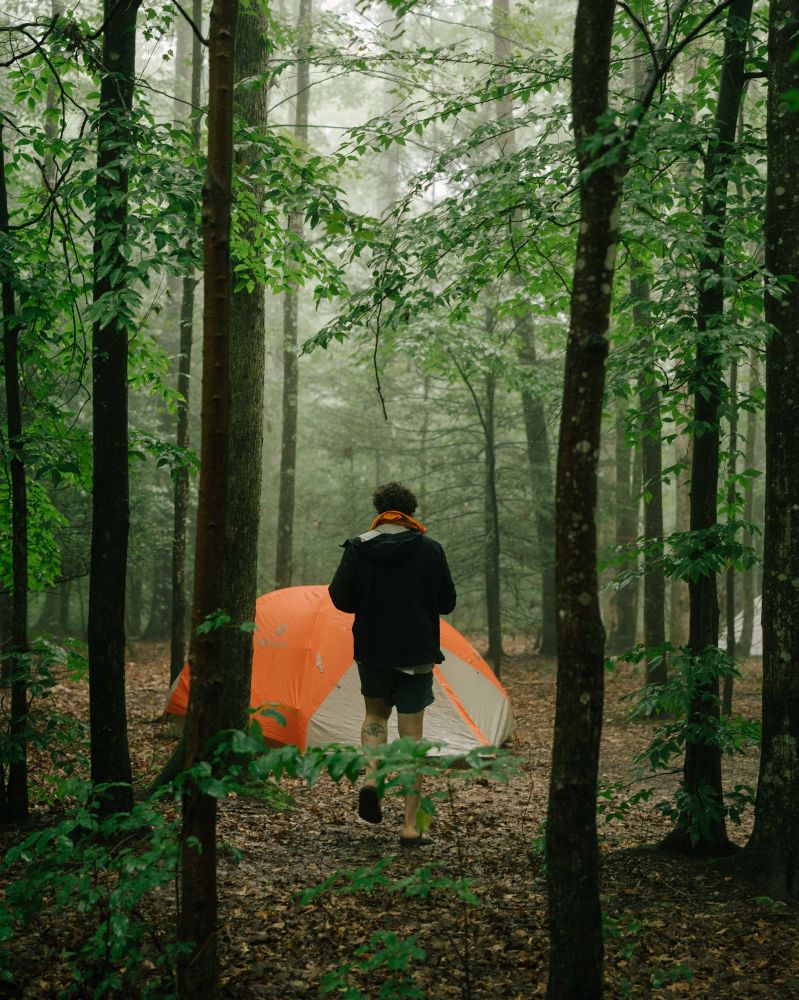 A man walks through a wet and rainy forest to his bright orange tent. 