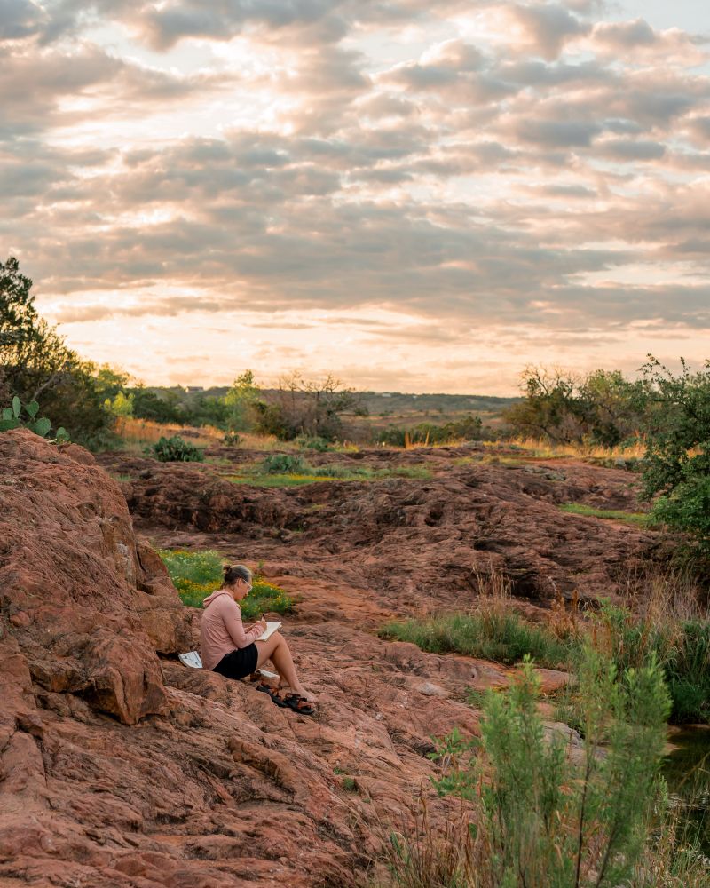 A women sits on a rocky foreground writing in her journal as the sun comes up. 