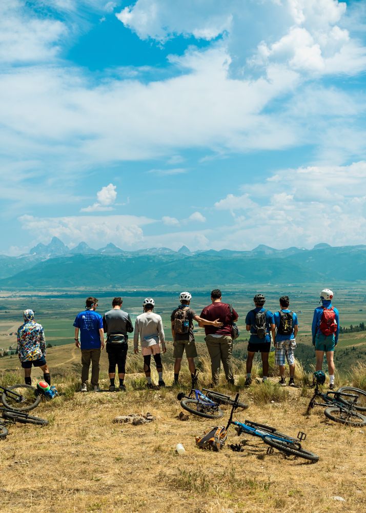 A group of men stand with their backs facing the camera looking off at the Tetons, with mountain bikes scattered in the foreground. 